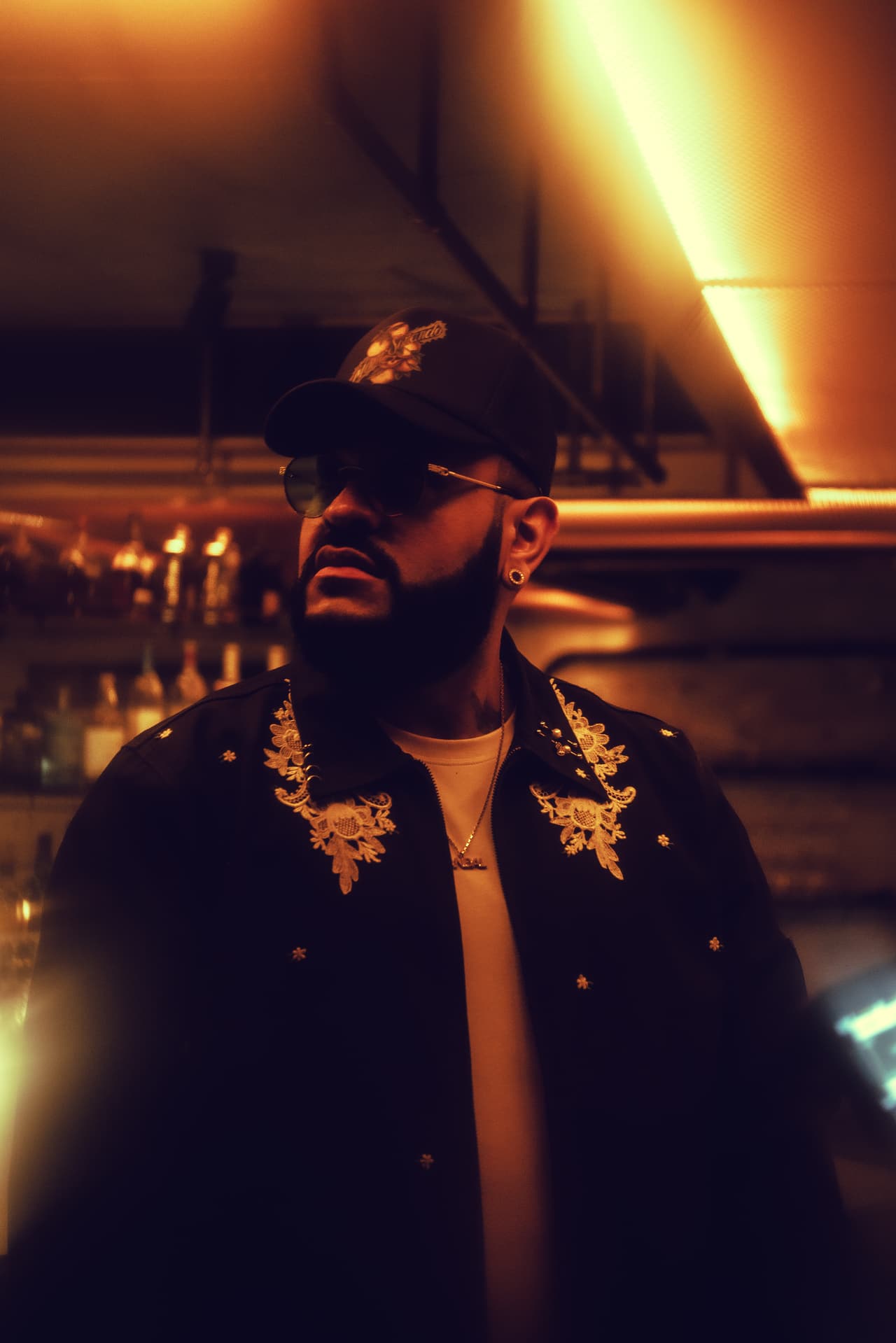A man at a bar under a gold-lit metal-mesh ceiling panel, backlit bottle shelves behind him, chin lifted