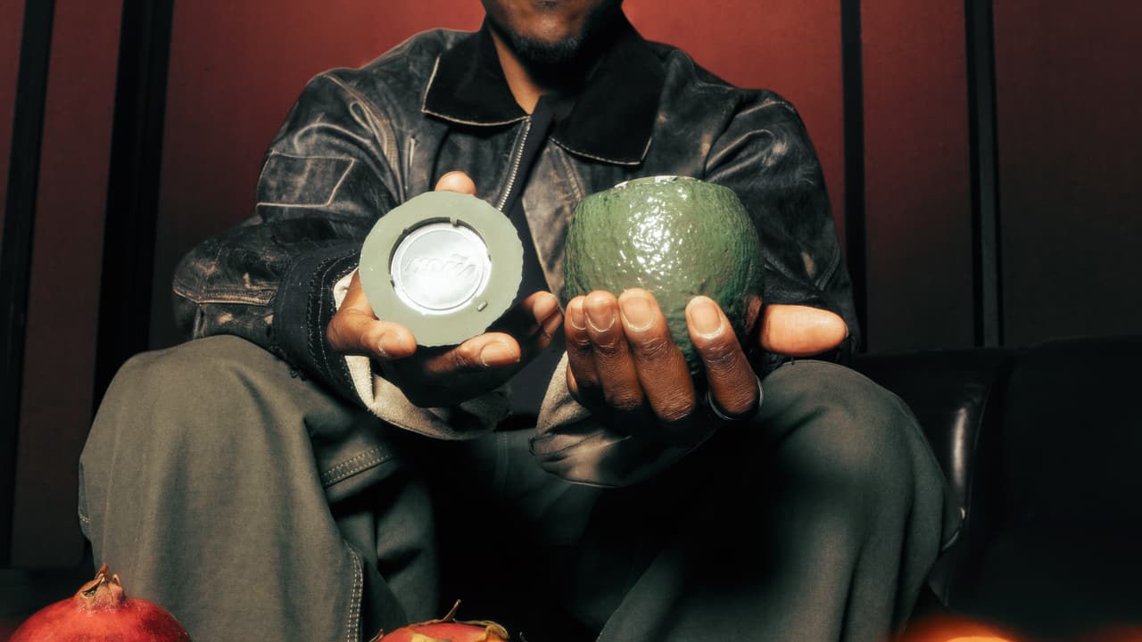 A man holding out a green ceramic avocado in one hand and its lid in the other, a silver MOÑA-embossed disc set into the lid