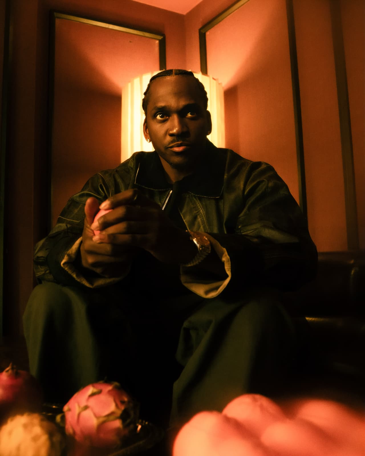 A man on a leather sectional looking straight down the lens, pomegranate and dragon fruit along the bottom edge, warm red light