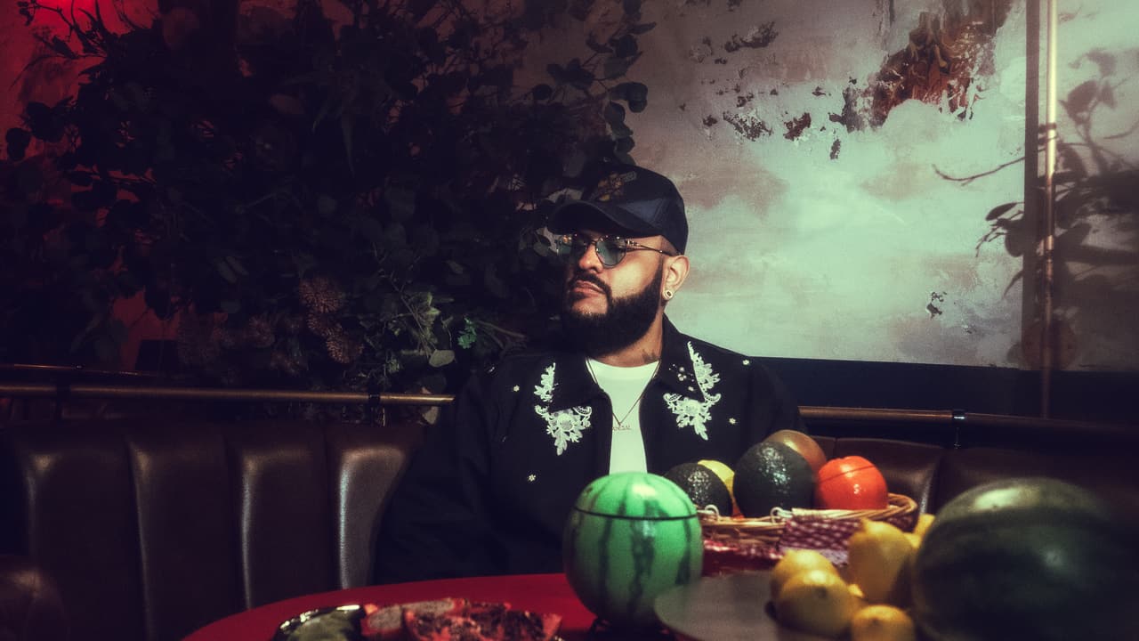 A man in a navy citrus-crest trucker and a white-lace-appliqué jacket behind a red-clothed table of melons and lemons, a red EXIT sign glowing at the edge of the frame