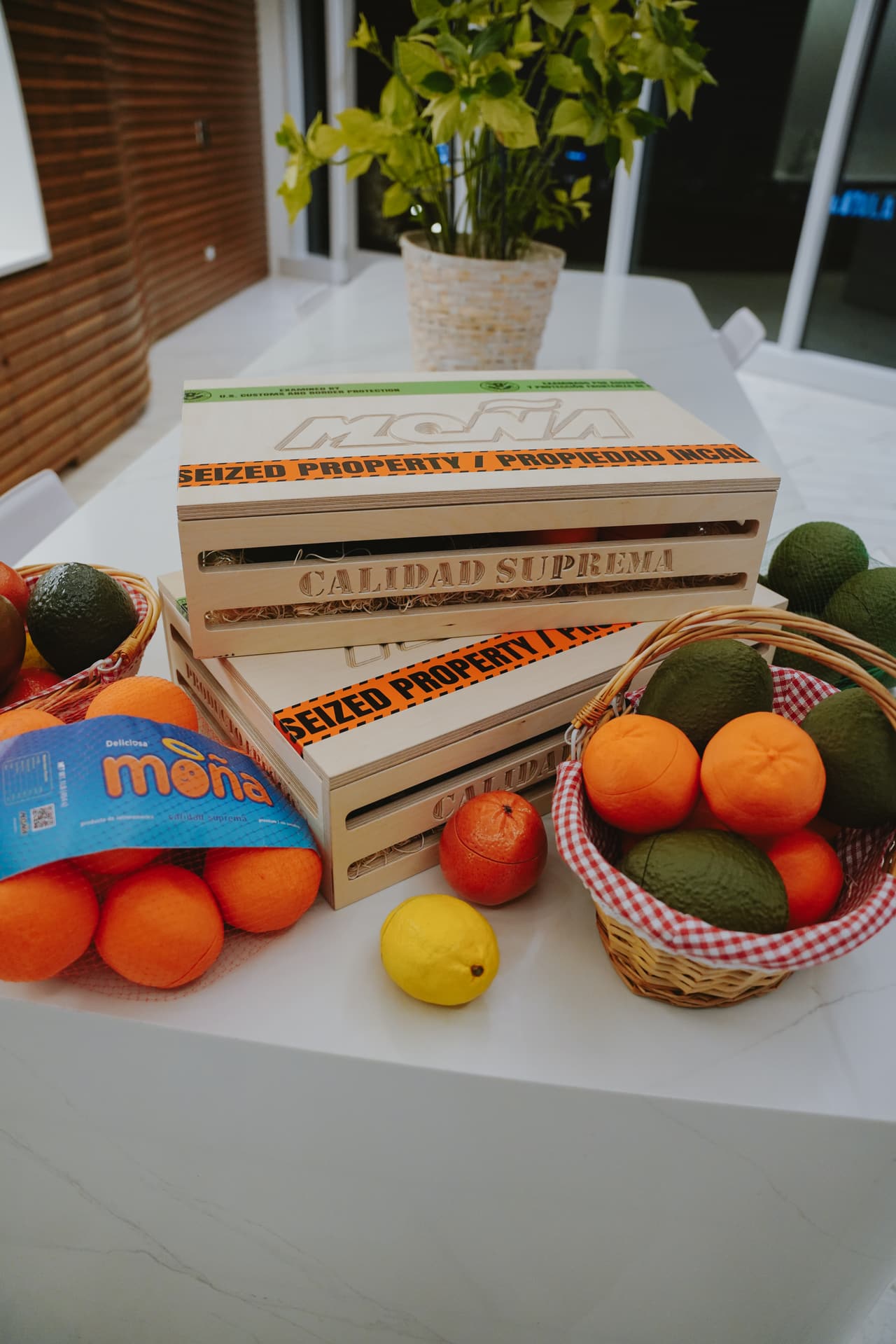 Stacked crates stamped 'CALIDAD SUPREMA' with fruit baskets, an orange net bag and a lemon, a plant behind