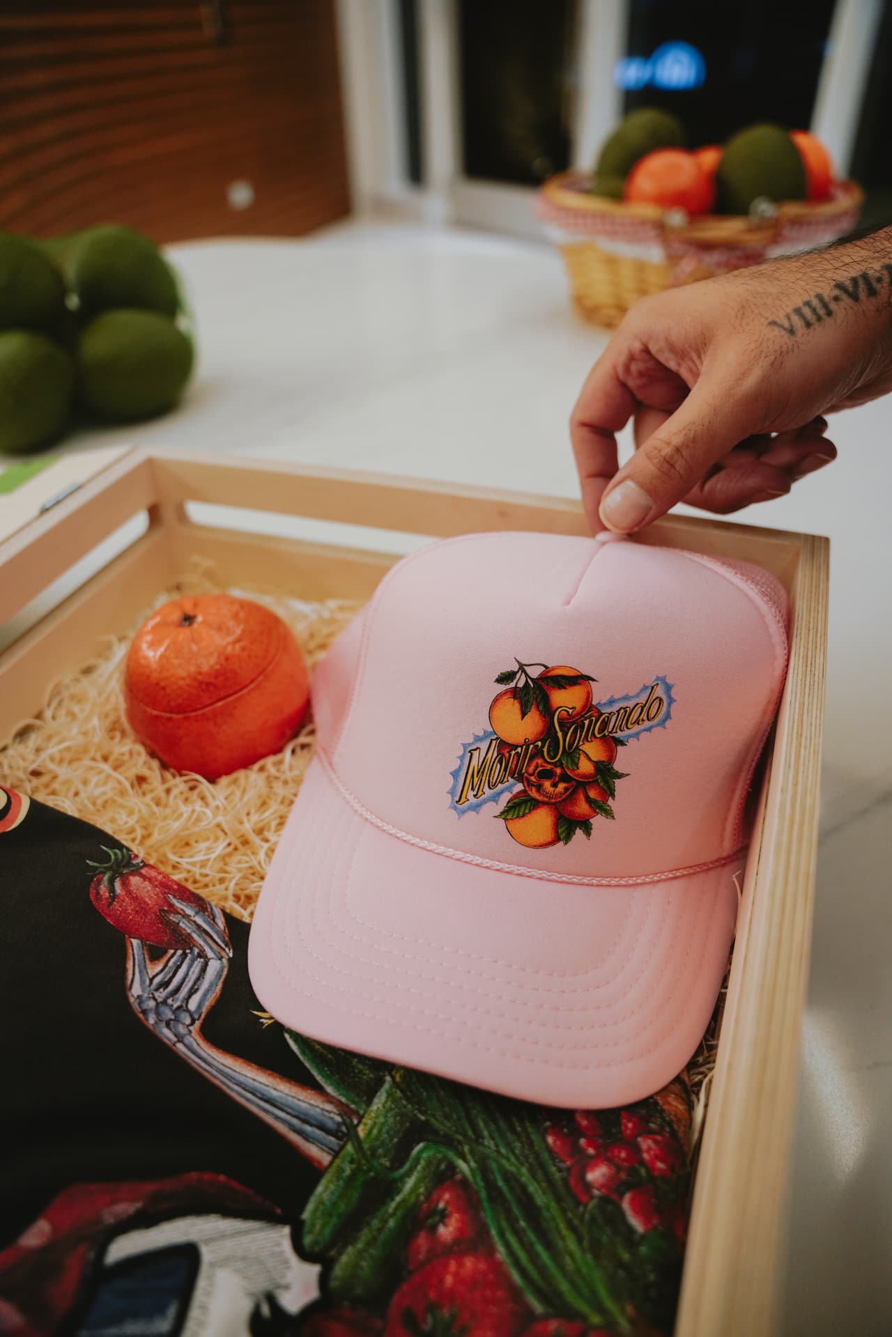 A hand lifting the pink trucker hat from a crate over a folded harvest tee
