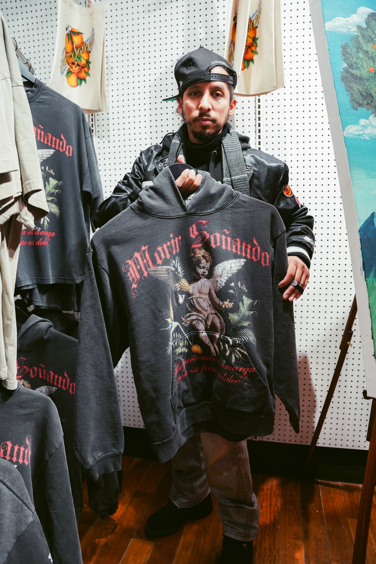 A guest in a satin jacket holding up the black cherub hoodie, more merch hanging on a pegboard behind