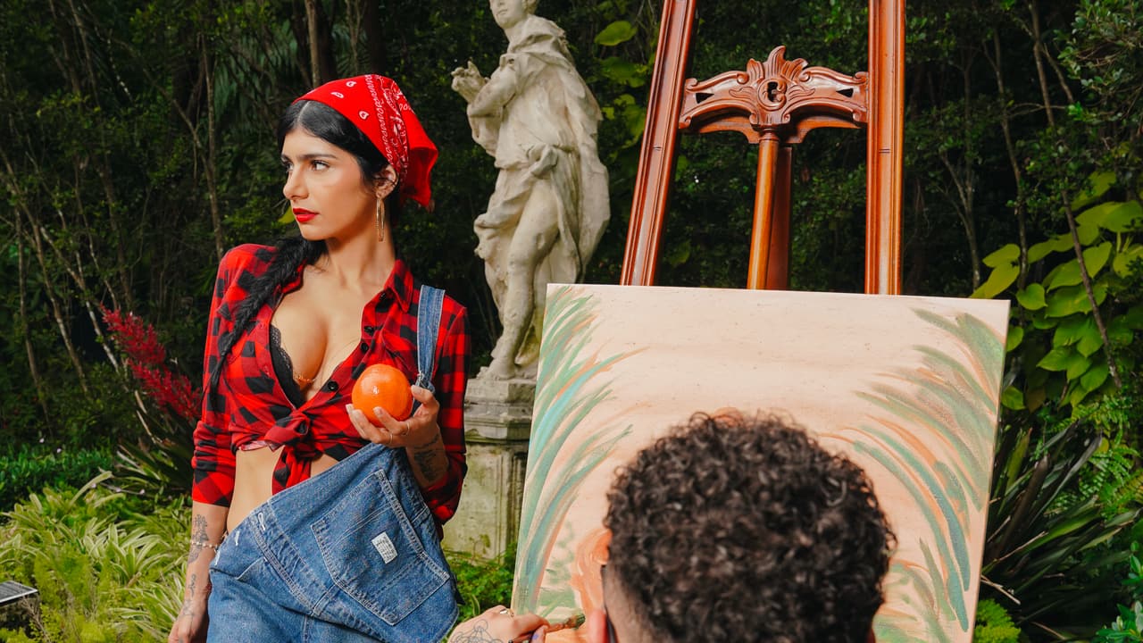 Over the shoulder of a man painting an orange-basket canvas at an easel in a garden, a woman in red boots holding a fruit basket beside a statue