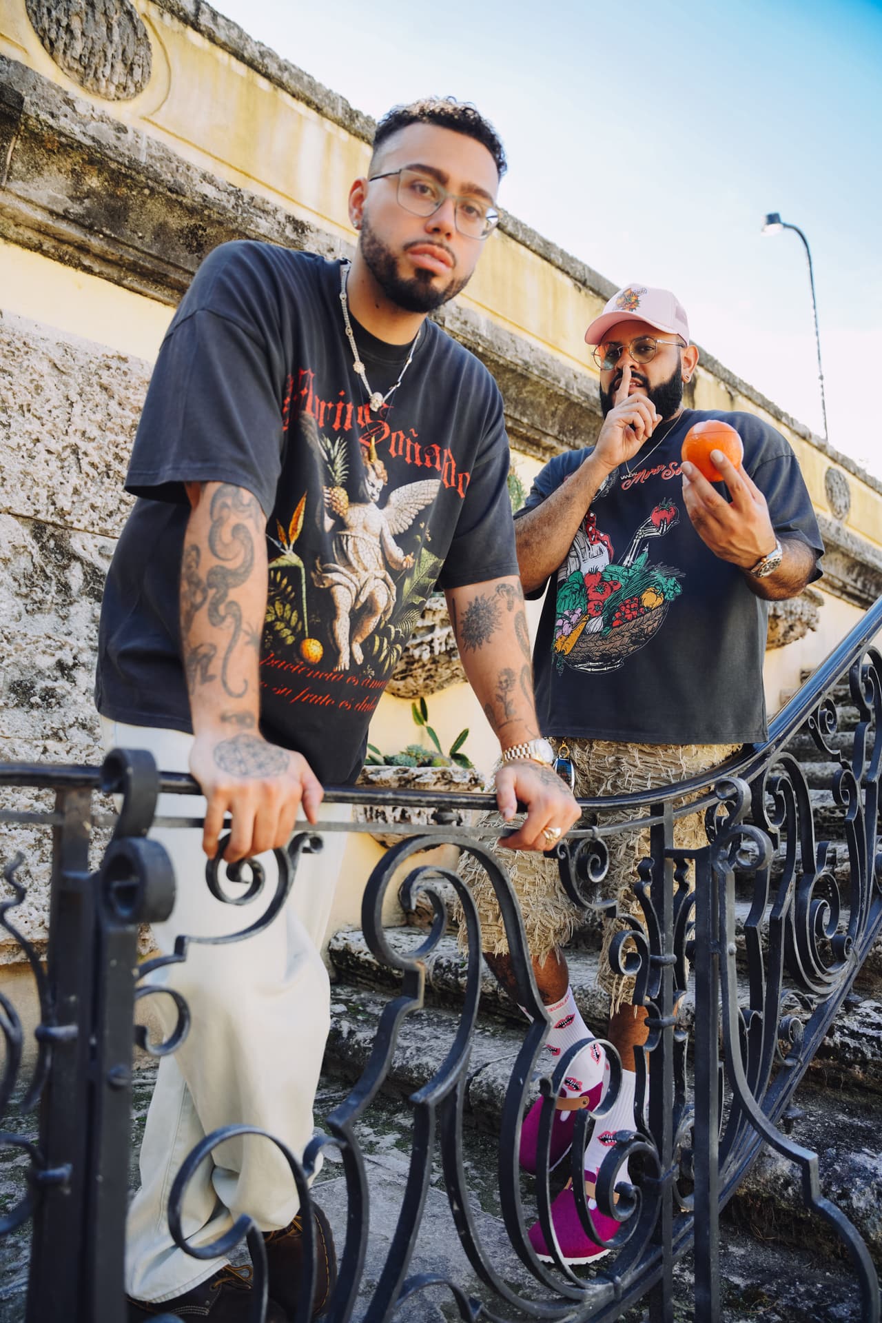 Two men at an iron railing, one leaning in wearing the cherub tee, the other making a quiet gesture while holding an orange