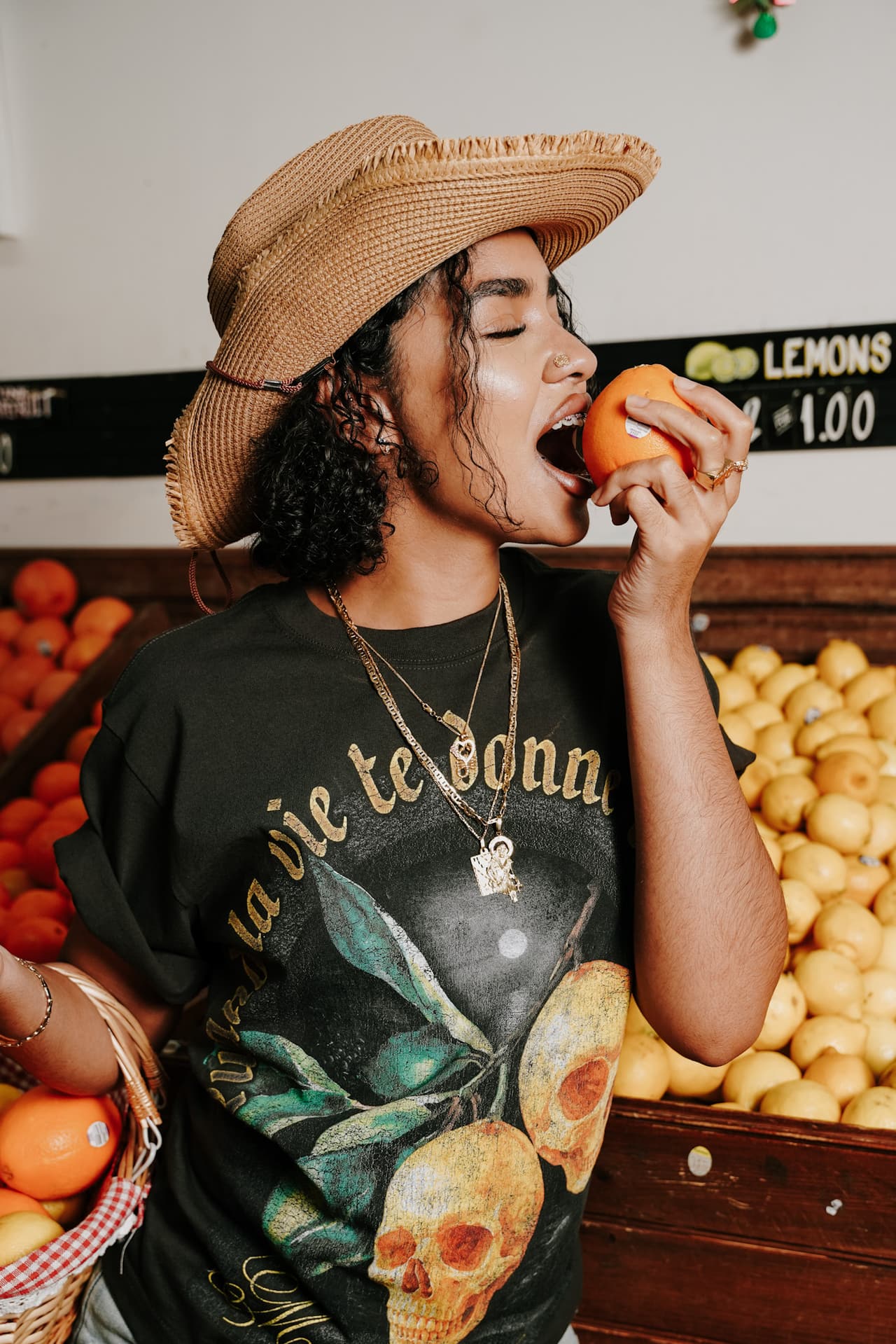 Close shot of the model biting into an orange, eyes closed, a 'LEMONS' sign and basket behind