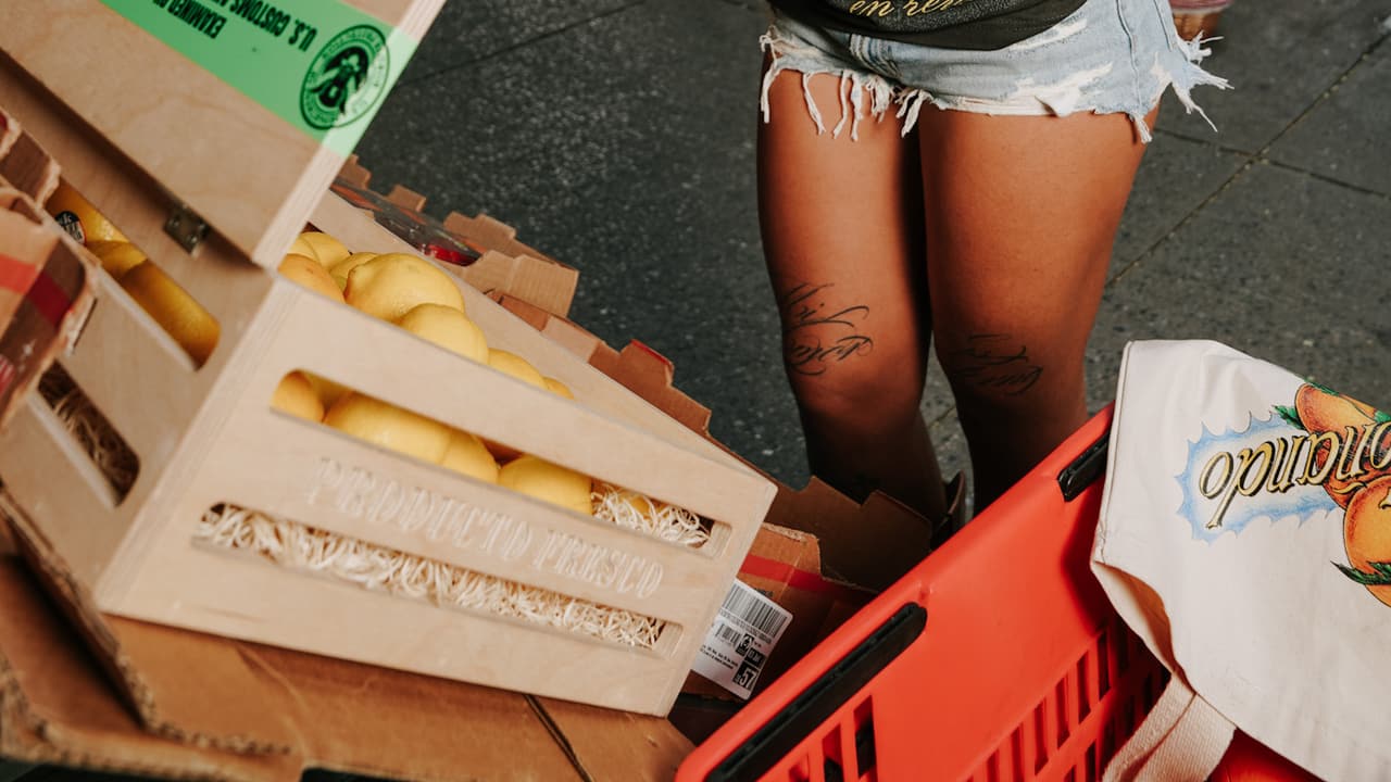 Model opening a wooden MOÑA crate stamped 'SEIZED PROPERTY' full of lemons on the sidewalk, a city street behind