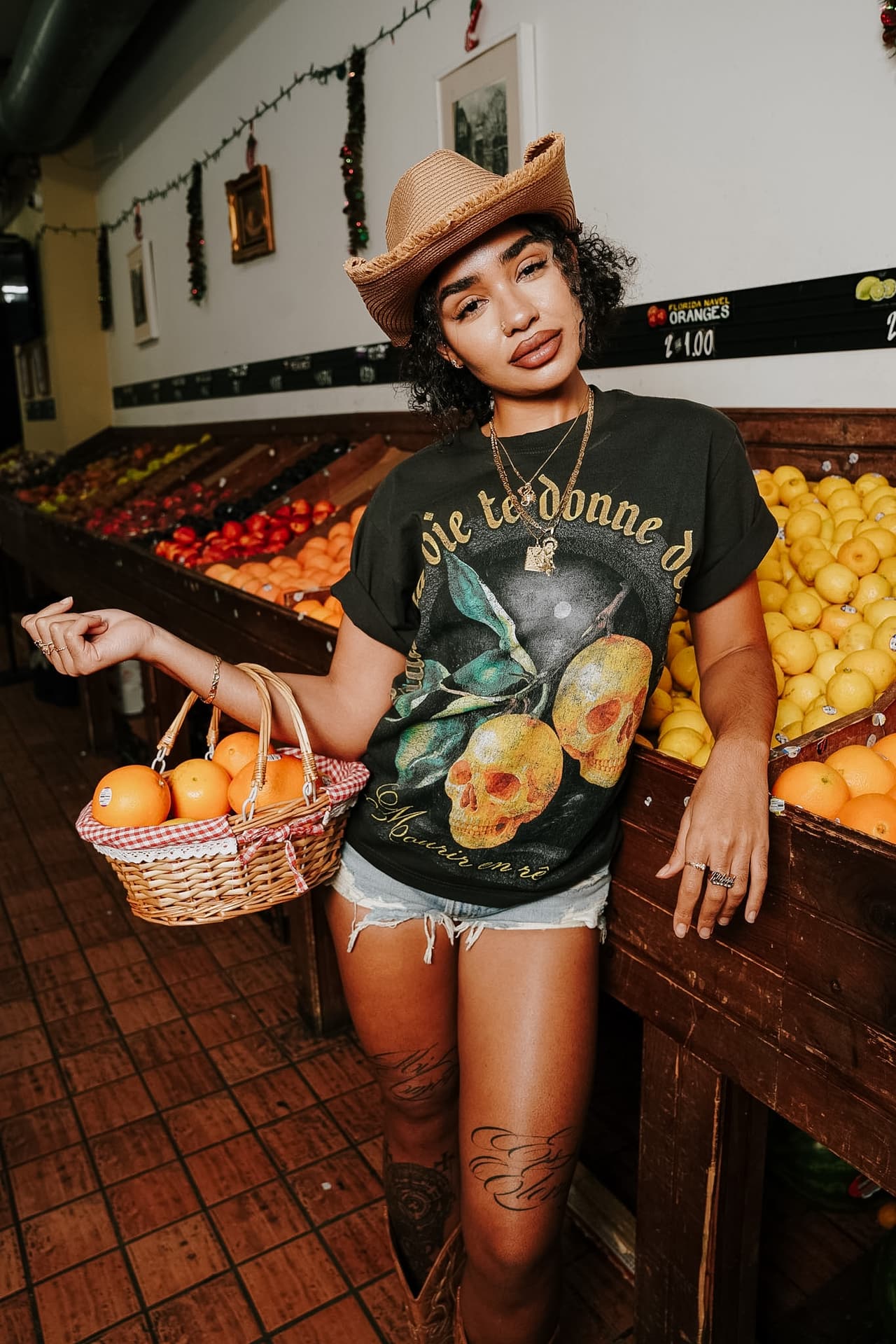 Model leaning on a crate of lemons inside the bodega, a basket in hand, garland lights overhead