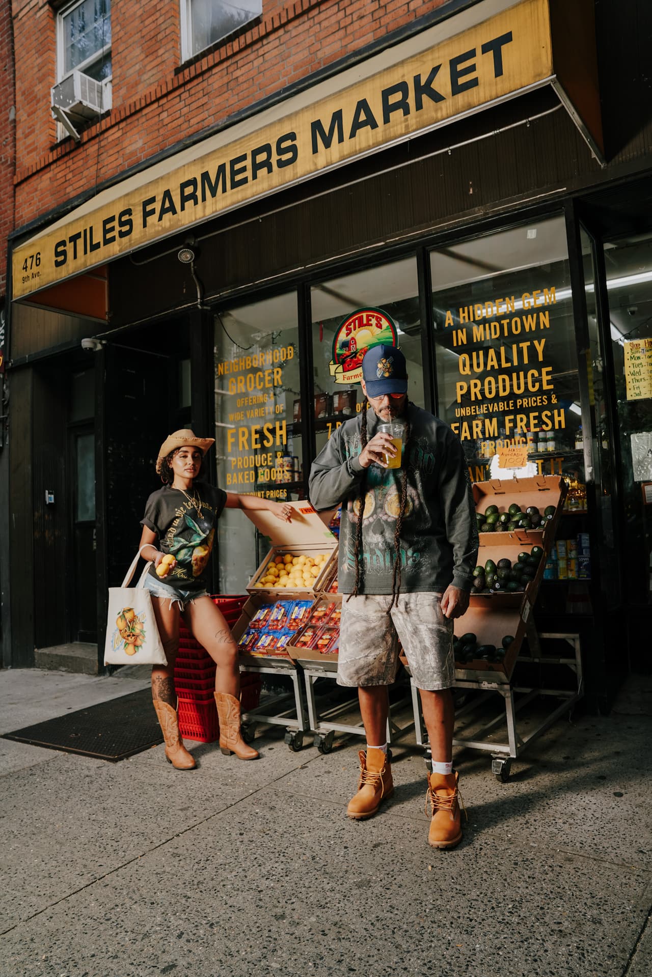 Model with a tote and a man in a long-sleeve tee sipping juice, both under the Stiles awning